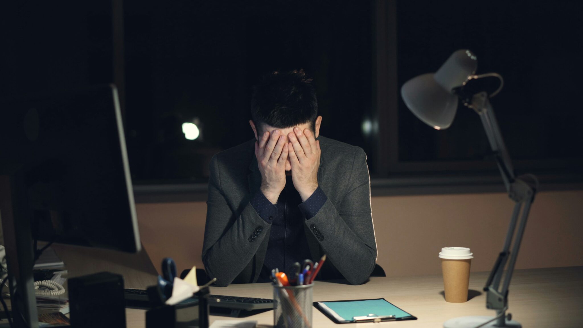 A stressed man sitting at a desk at night with his face in his hands, surrounded by office equipment and paperwork.