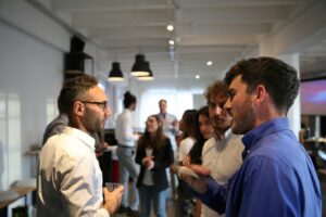 A group of men in button-down shirts having a conversation at a networking event, with other attendees mingling in a modern, well-lit loft space behind them.