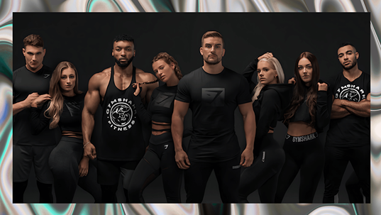 Group of eight fitness models posing together in all-black Gymshark activewear against a dark studio backdrop.