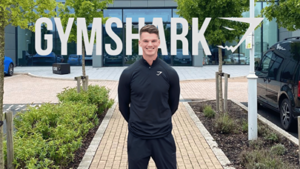 Man in black Gymshark quarter-zip standing outside an office building with a large "Gymshark" sign overhead.